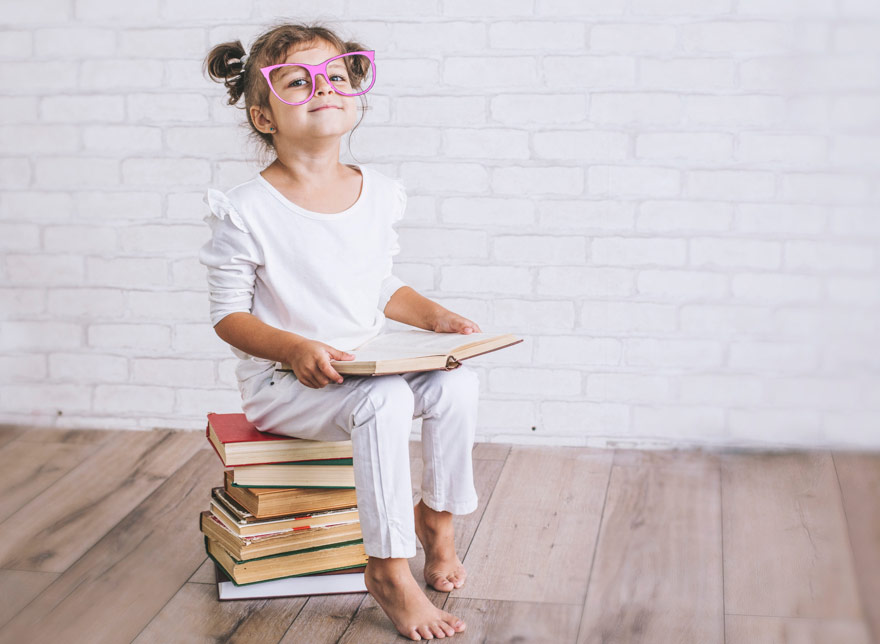 Little girl sitting on a pile of books with oversized pink glasses.